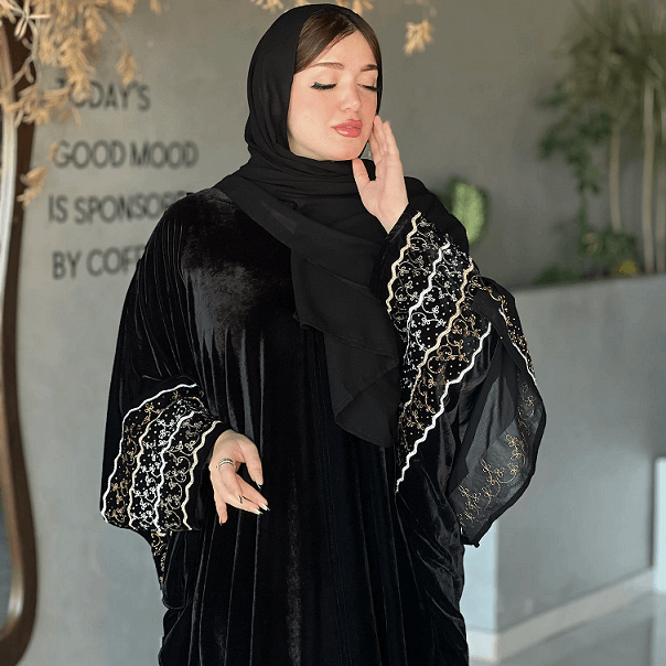 Woman wearing a black abaya with white embroidery in a room with plants and a motivational quote on the wall.