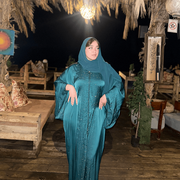 Woman in a teal dress standing in an outdoor setting with wooden furniture and plants.