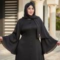 Woman wearing a black abaya standing outdoors with a building and fence in the background.